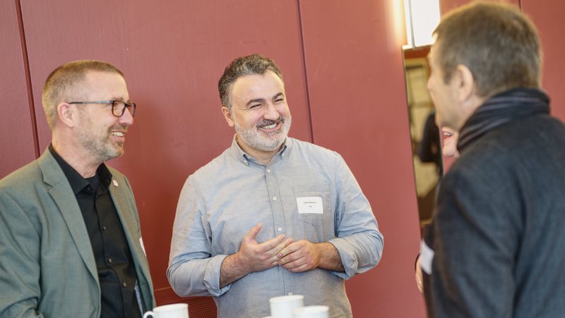 Das Foto zeigt eine Gruppe von Menschen, die sich in einer entspannten Atmosphäre unterhalten. Drei Männer stehen um einen Stehtisch mit Kaffeetassen darauf. Sie lachen und scheinen sich in einer freundlichen und professionellen Umgebung zu befinden, möglicherweise bei einer Netzwerkveranstaltung oder Konferenz. Der Hintergrund ist eine rotbraune Wand mit einer Lichtquelle an der Seite.