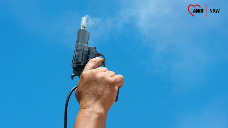 Auf dem Foto ist eine Hand zu sehen, die eine Startpistole in den Himmel hält. Die Pistole scheint gerade abgefeuert worden zu sein, da Rauch aus dem Lauf austritt. Der Hintergrund besteht aus einem klaren blauen Himmel. Außerdem ist oben rechts das Logo der AWO NRW zu sehen.