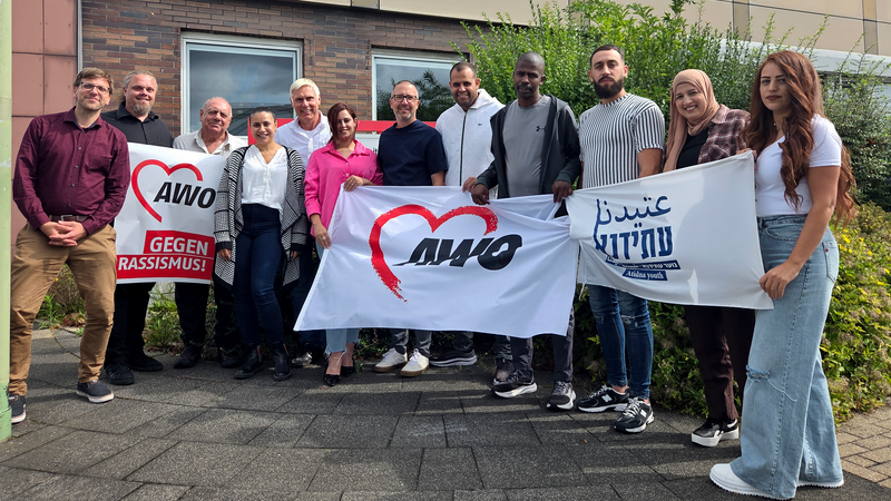 Auf dem Foto ist eine Gruppe von etwa einem Dutzend Personen zu sehen, die vor einem Gebäude stehen. Sie halten drei Banner hoch: Ein weißes Banner mit einem roten Herz und dem Slogan "AWO" in Rot. Ein weiteres weißes Banner mit einem roten Herz und dem Text "AWO GEGEN RASSISMUS!" in Schwarz. Ein weißes Banner mit Text in einer ausländischen Schrift. Die meisten Personen auf dem Foto tragen lässige Kleidung. Die Stimmung wirkt positiv und die Personen schauen freundlich in die Kamera.